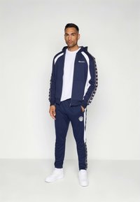 Fato de treino azul-marinho com detalhes brancos, casaco com fecho de correr e logótipo. Calças com riscas laterais e detalhe de logótipo. Usado com ténis brancos.