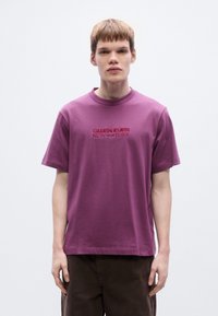 Μωβ βαμβακερό t-shirt με στρογγυλή λαιμόκοψη και κοντά μανίκια. Διαθέτει τη ρίγα "CALVIN KLEIN NEW YORK" κεντημένη με κόκκινο χρώμα στο μπροστινό μέρος.