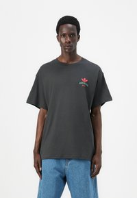 T-shirt oversize grigio scuro in cotone, con un piccolo logo Adidas rosso e verde sul petto. Maniche corte, vestibilità rilassata.