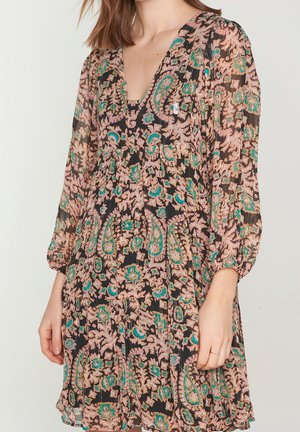 Femme portant une robe à manches longues avec un motif floral cachemire rose, sarcelle et noir et un décolleté en V, debout devant un fond uni.