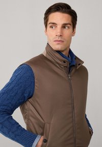 Gilet imbottito marrone con colletto alto, zip frontale e tasche laterali, indossato sopra un maglione blu a maglia a treccia con una camicia a scacchi sotto.