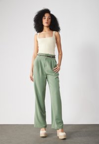 Lys beige ribbet tanktop, høyt liv grønne bukser med folder, og beige plattform espadrillos. Minimalistisk design, myk tekstur.