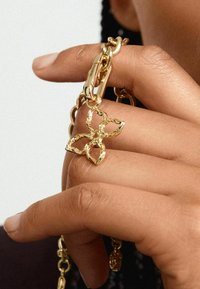 Guldmetallkedja med en texturerad fjärilscharm som hålls av en hand. Charmet har en öppen design och är fäst vid en kläpp.