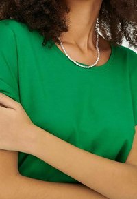 T-shirt di cotone verde con collo a giro e maniche corte arrotolate. Presenta una trama liscia ed è abbinato a una collana di perle.