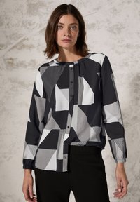 Blouse à motif noir et blanc avec des formes géométriques, devant à boutons, manches longues avec poignets élastiques, fabriquée en tissu léger.