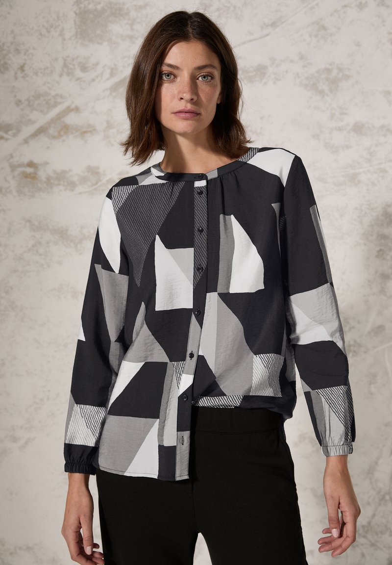Blouse à motif noir et blanc avec des formes géométriques, devant à boutons, manches longues avec poignets élastiques, fabriquée en tissu léger.