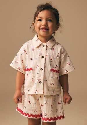 Lachend jong meisje in een crème outfit met rood- en blauwbloemige patronen en rode bies op zakken en korte broek tegen een beige achtergrond.