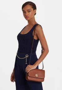Borsa a tracolla in pelle marrone con una chiusura a logo dorato. Sfondo neutro, modella in top di maglia blu navy con un accentuato cinturino a catena.
