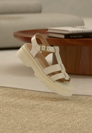 Bež sandal s debelejšim podplatom, tremi širokimi trakovi in zlatim zaponkom, postavljen na teksturirano bež preprogo blizu lesenega kavčnega mizice s knjigami.