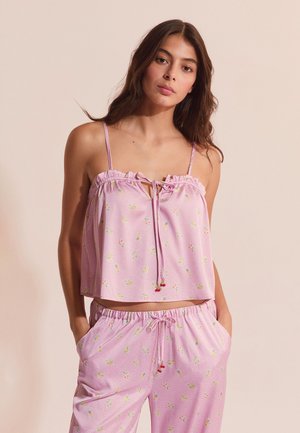 Junge Frau trägt ein ärmelloses, rosa, geblümtes Oberteil und passende Hosen mit Tunnelzug, steht mit den Händen in den Taschen vor einem schlichten Hintergrund.