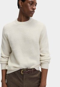 Helles, geripptes Sweatshirt mit Rundhalsausschnitt und umgeschlagenen Bündchen, kombiniert mit braunen Cordhosen und einem dunklen Gürtel mit einer goldenen Schnalle.