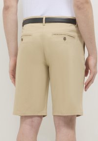 Beige Herren Shorts mit zwei geknöpften Gesäßtaschen und einem schwarzen Gürtel, getragen mit einem weißen, in die Hose gesteckten Hemd, von hinten gezeigt.