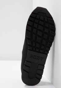 Semelle de sneaker noire avec un motif en caoutchouc texturé, arborant un logo "DKNY" bien visible et une légère élévation au niveau du talon.