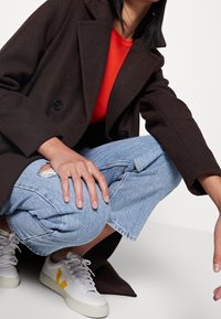 Hnědý dvouřadový kabát, červená košile, světle modré džíny s otřesy a bílé tenisky se žlutými akcenty. Zaměřeno na detaily látky a střihu.