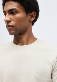 Άνδρας με κοντά σγουρά μαλλιά που φοράει μια μονόχρωμη ανοιχτό μπεζ μπλούζα με στρογγυλή λαιμόκοψη, κοιτώντας προς τα αριστερά μπροστά σε ένα ανοιχτό γκρι φόντο.