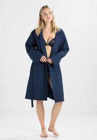 Robe longue bleu marine en matériau texturé, avec de longues manches, une ceinture à la taille et deux poches avant. Design ouvert avec une coupe décontractée.