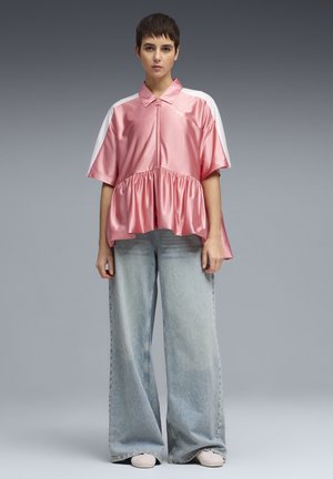 Persona de pie con una blusa suelta de satén rosa de manga corta con rayas blancas y vaqueros azules desgastados de pierna ancha.