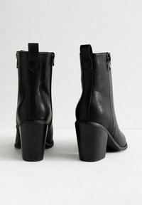 Botas de tobillo de cuero negro con puntera redondeada, cremalleras laterales y un tacón grueso. Presentan una textura suave y detalles de costura sutiles.