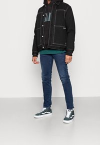 Giacca di jeans nera con cuciture bianche, indossata sopra un hoodie nero, abbinata a jeans slim-fit blu scuro e sneakers alte verde.