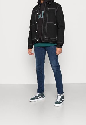 Giacca di jeans nera con cuciture bianche, indossata sopra un hoodie nero, abbinata a jeans slim-fit blu scuro e sneakers alte verde.
