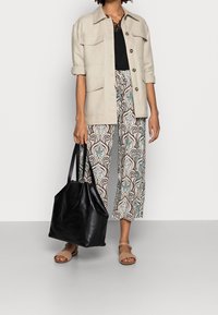 Veste utilitaire beige à manches retroussables portée sur un haut noir, pantalon large à motifs bleu et marron, sac cabas noir, et sandales couleur tan.