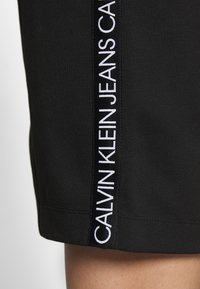 Czarne szorty z bocznym paskiem z napisem "CALVIN KLEIN JEANS" w wyrazistym białym kolorze. Gładki materiał, dopasowany krój i czysta krawędź.