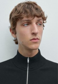 Zwarte zip-up trui met hoge kraag, voorzien van een zilveren rits. De stof lijkt gebreid en het ontwerp is minimalistisch.