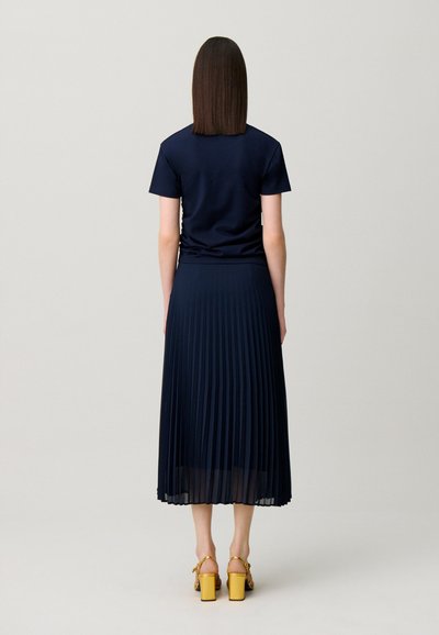 Donna che indossa una maglia blu navy a maniche corte, gonna midi plissettata abbinata e sandali gialli con tacco alto, in piedi di spalle.