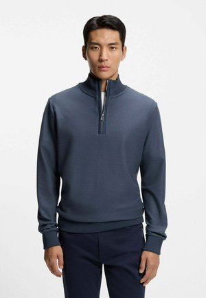 Homme portant un pull à demi-fermeture éclair bleu marine et un pantalon foncé, debout devant un fond clair uni.