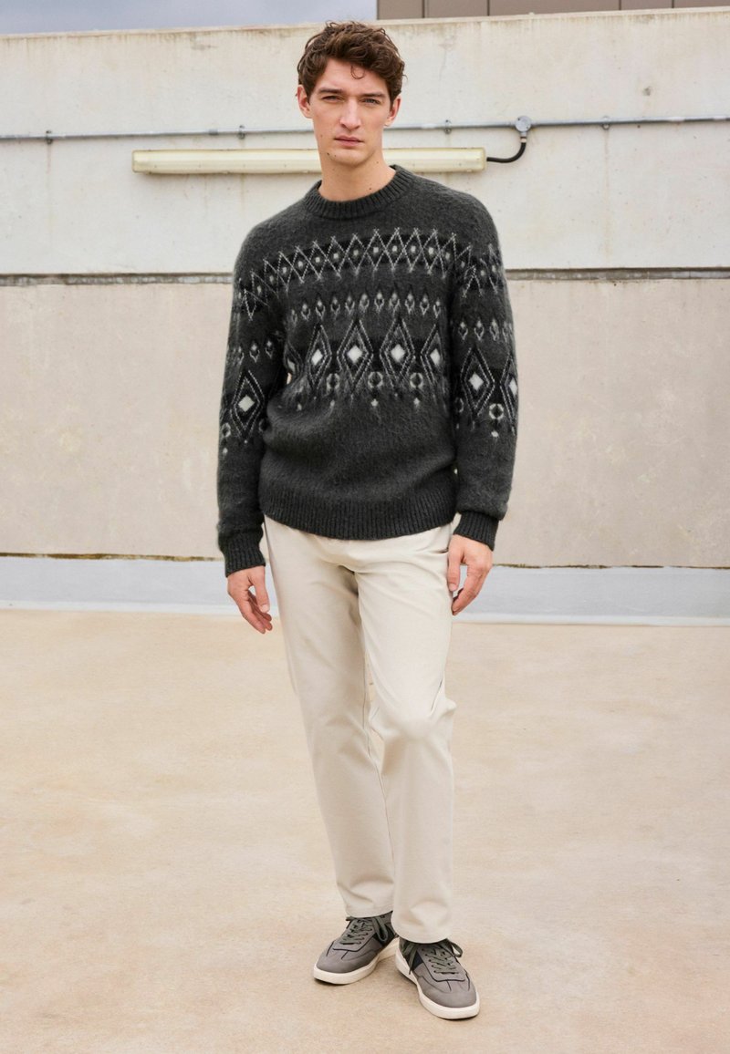 Mørkegrå strikket sweater med et geometrisk hvidt mønster, parret med lysebeige bukser og grå sneakers, som viser et afslappet outfit.
