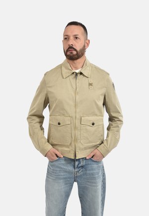 Hombre con chaqueta beige con cremallera y dos bolsillos delanteros y vaqueros azul claro, de pie con las manos en los bolsillos frente a un fondo liso.