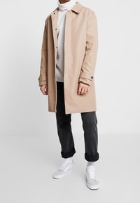 Beżowy trencz z kołnierzem, ozdobiony guzikami, z podwiniętymi rękawami, zestawiony z szarym swetrem, czarnymi spodniami i białymi trampkami.