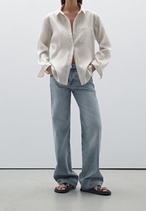 Light beige button-up shirt with long sleeves and a chest pocket, paired with loose light blue jeans and black sandals. Smooth fabric texture.