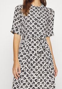 Šaty s leopardím potiskem v černobílé, krátké rukávy, zapínání na knoflíky, látkový pásek v pase, lehký materiál, splývavý design.