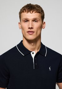 Marinblå polo skjorta med vit krage, korta ärmar och knappslå med tre knappar. Har en liten logotyp på vänster bröst. Slät yta.