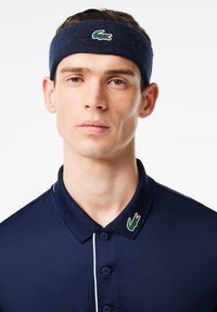 Ναυτικό μπλε βαμβακερό πουκάμισο πόλο με κουμπιά, λευκά αξεσουάρ και πράσινο λογότυπο κροκόδειλου στον γιακά; σε συνδυασμό με μια αντίστοιχη ναυτική κορδέλα κεφαλής.