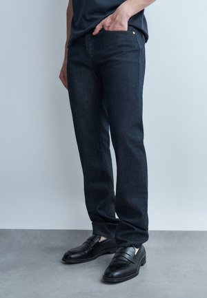 Osoba ubrana w ciemnoniebieskie dżinsy, z ręką w kieszeni i czarne skórzane mokasyny, stojąca na szarej podłodze na tle jasnej ściany.