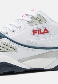 Valge Fila toss, millel on punane logo, valged paelad, hallid ja tumesinised aktsendid ning tekstureeritud kummist tald valge tausta peal.