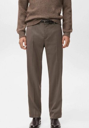 Mand iført brun strikket sweater, taupe plisserede bukser med sort bælte og mørkebrune lædersko, stående mod en ensfarvet baggrund.