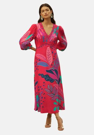 Žena stojaca s rukami na bokoch, oblečená v červenom maxi šatách s veľkými vzormi listov v ružovej, fialovej a zelenej farbe a balónovými rukávmi.