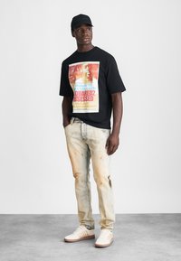 Μαύρο μπλουζάκι με πολύχρωμο γραφικό τύπωμα μπροστά. Ανοιχτό μπλε τζιν με φθαρμένες λεπτομέρειες. Λευκά αθλητικά παπούτσια με μπεζ λεπτομέρειες.