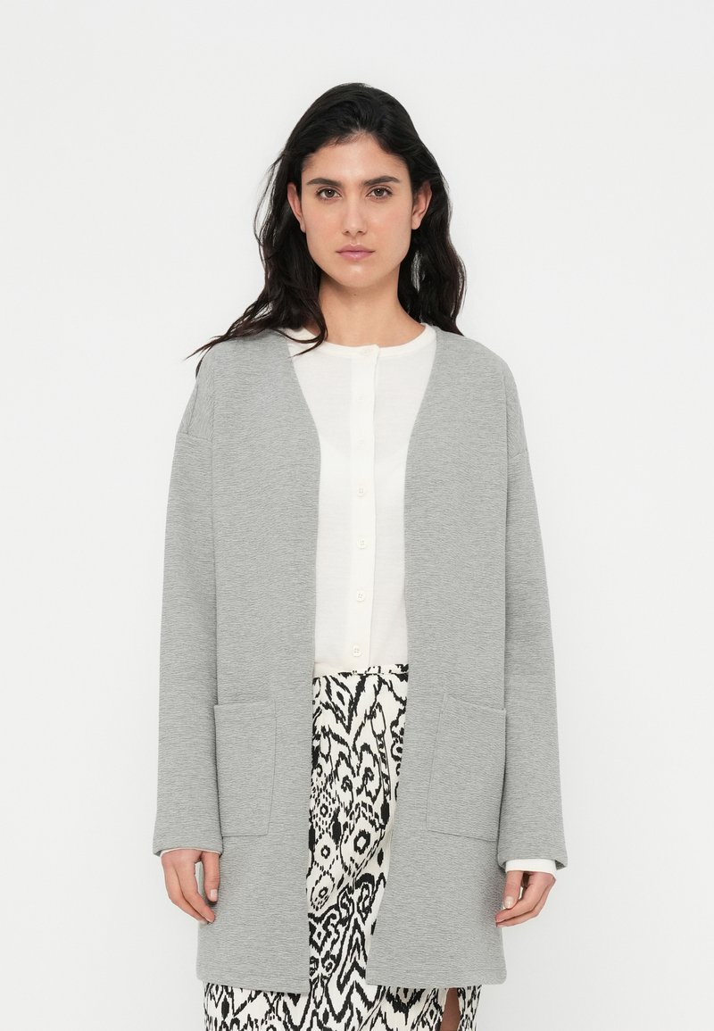 Γκρι ζακέτα με μακριά μανίκια, ανοικτή μπροστά, δύο εμπρόσθιες τσέπες, φορεμένη πάνω από λευκό πουκάμισο με κουμπιά και φούστα με ασπρόμαυρο μοτίβο.