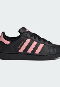 Crna kožna tenisica s ružičastim prugama i naglascima. Ima gumeni potplat, vezice i teksturirani vrh prsta. Vidljivo je "SUPERSTAR" obilježje.