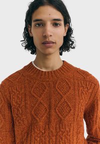 Jeune personne ayant des cheveux bouclés de longueur moyenne et de couleur sombre, portant un pull en tricot torsadé orange, se tournant vers l'avant contre un fond uni.