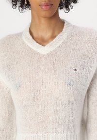 Lightweight, sheer white knit sweater with a v-neck, featuring a small logo detail on the left side. Soft texture and loose fit.