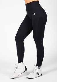 Black high-waisted leggings with a smooth texture, side panel details, and a small white logo on the hip. Worn with white athletic shoes.