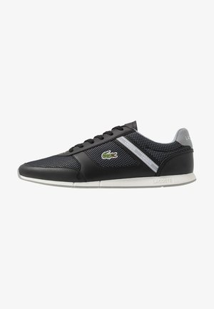 Zapatilla negra Lacoste con paneles de malla, suela blanca, logo de cocodrilo verde y detalles grises, vista de lado.