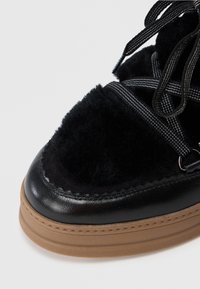 Chaussure en cuir noir et en shearling avec une épaisse semelle en caoutchouc marron et des lacets noirs, ornée de coutures blanches.