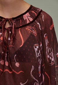 Bordeaux gennemsigtig bluse med en prikket tekstur, der har en velourkrave og snøre, prydet med legende mønstre af drikkevarer og kirsebær.