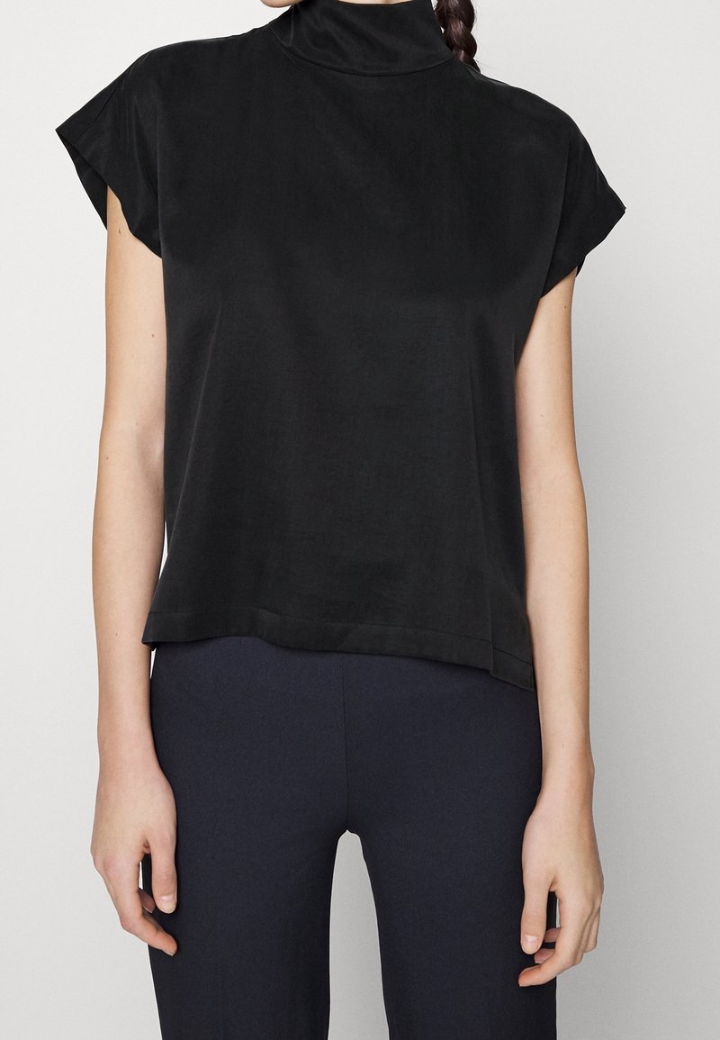 Haut noir à manches courtes avec un col montant, texture lisse, coupe décontractée et fentes sur les côtés, associé à un pantalon foncé ajusté.